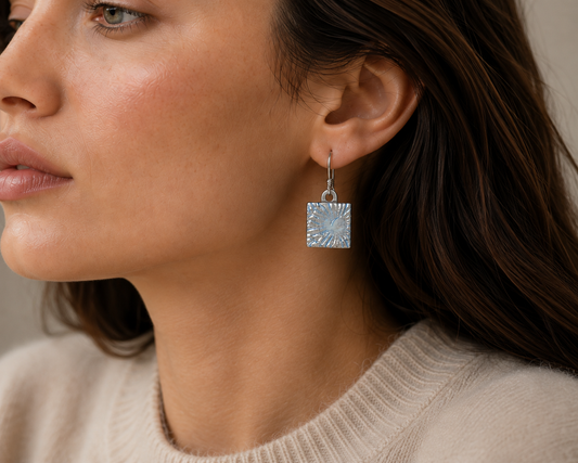 A close-up of a woman wearing a silver square drop earring with a textured sunburst pattern, paired with a beige knit sweater.