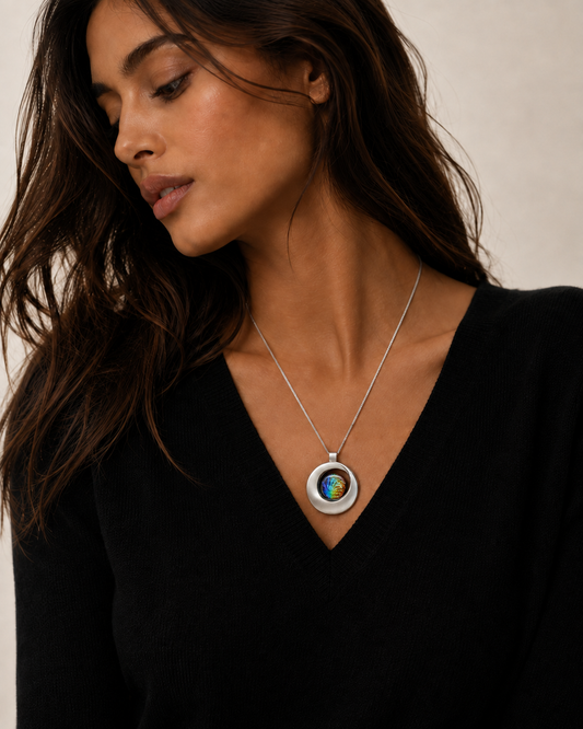 A woman wearing a silver pendant necklace with a circular, colorful inlay, styled with a black V-neck top against a neutral beige background.