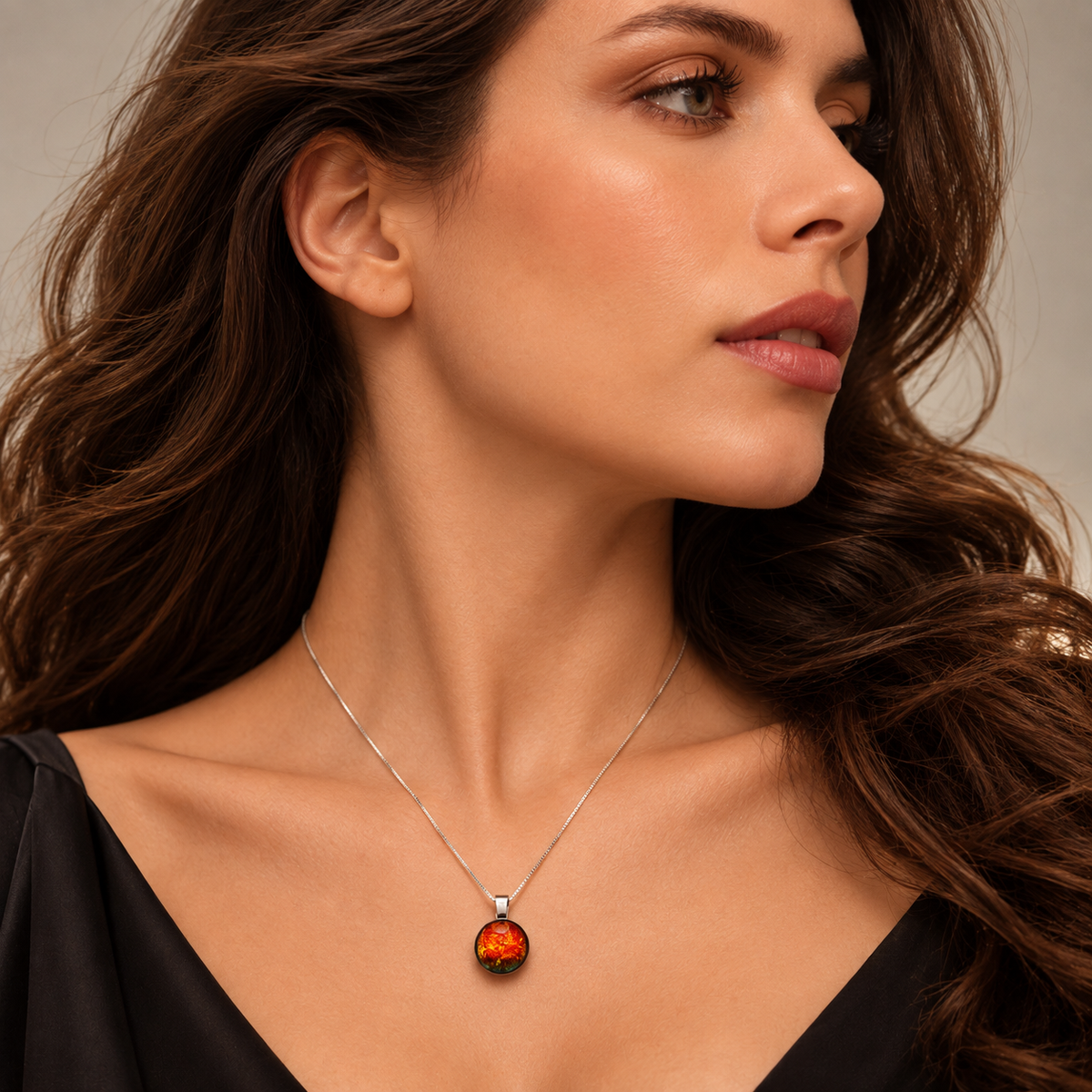 A close-up of a woman wearing a delicate silver chain necklace with a round pendant featuring a textured orange and red pattern, set against a soft beige background.