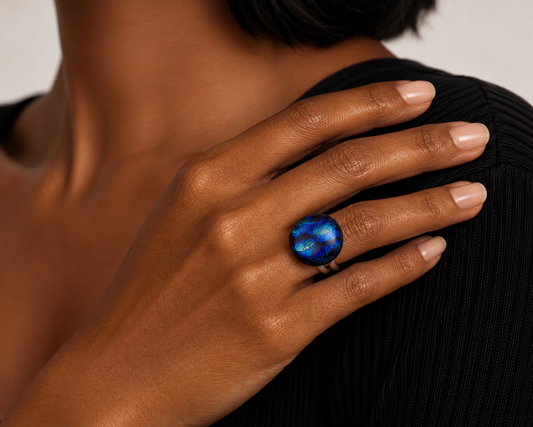 A close-up of a person's hand wearing a ring with a dark blue, swirling galaxy-like stone on a black ribbed top against a light neutral background.