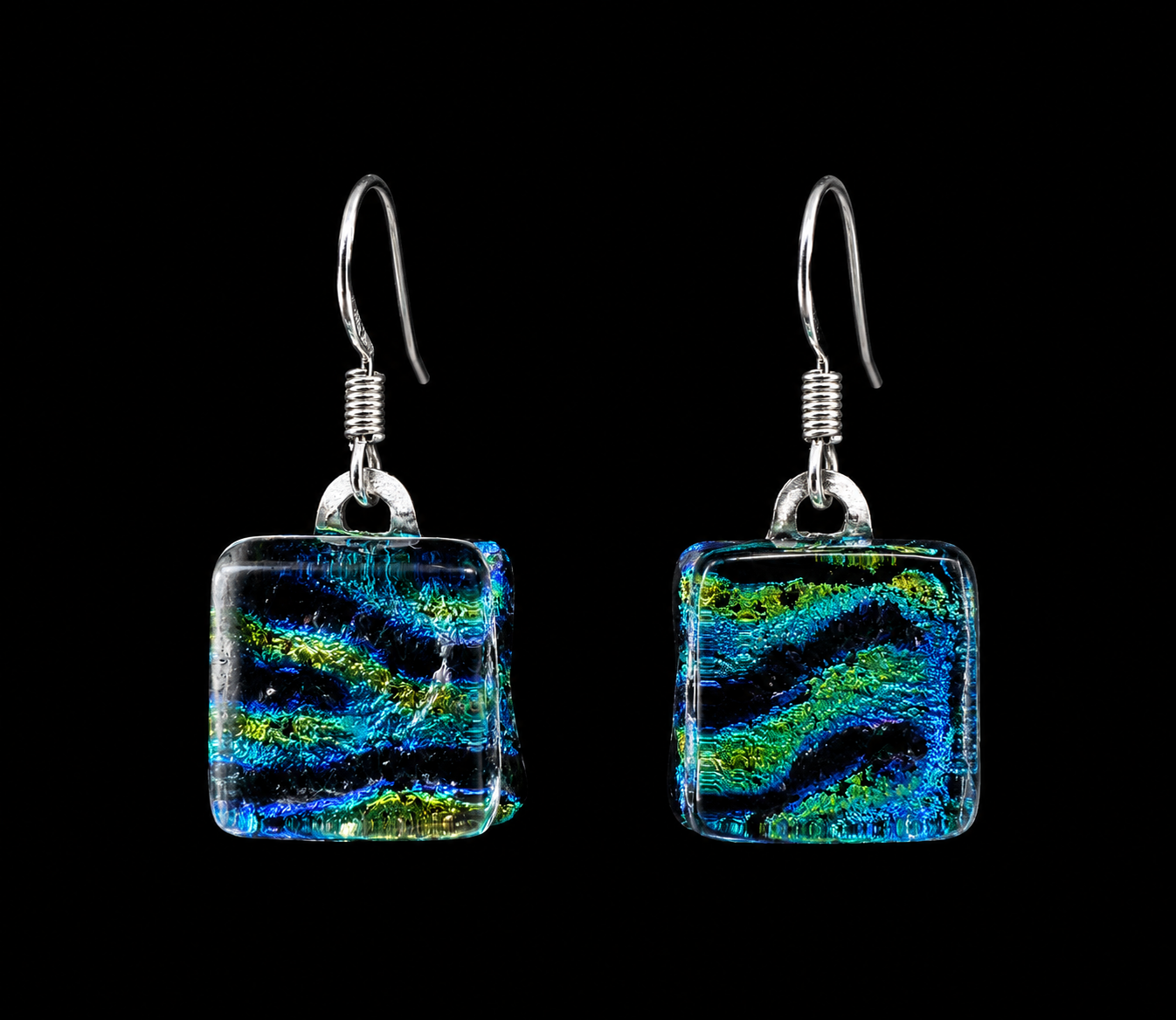 A pair of square glass drop earrings with a vibrant blue, green, and black swirled pattern, suspended from silver fishhook wires against a solid black background.