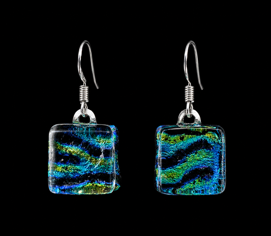 A pair of square glass drop earrings with a vibrant blue, green, and black swirled pattern, suspended from silver fishhook wires against a solid black background.