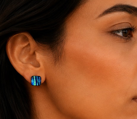 A close-up of a woman's ear wearing a small square stud earring with a vibrant blue iridescent, opal-like surface.