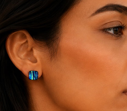 A close-up of a woman's ear wearing a small square stud earring with a vibrant blue iridescent, opal-like surface.