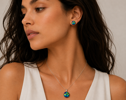 A close-up of a woman wearing a silver necklace and matching stud earring, both featuring a colorful round pendant or stud with a green, blue, and orange pattern, set against a plain light background.