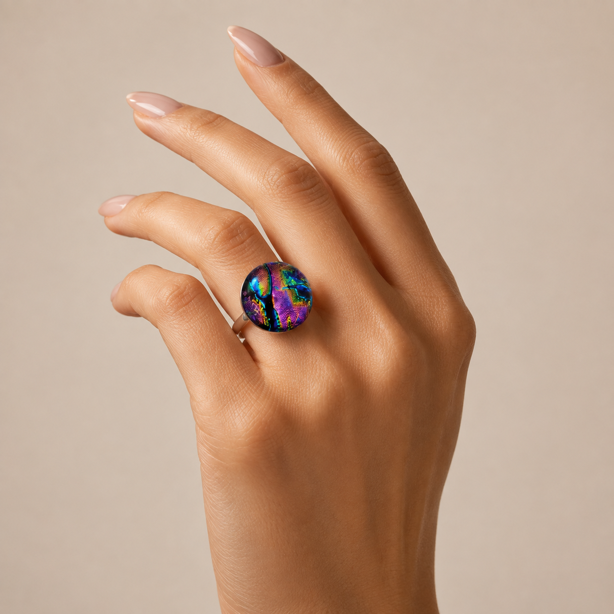 A close-up of a hand wearing a round iridescent ring with a multicolor opal-like pattern in blue, purple, green, and pink on a beige background.