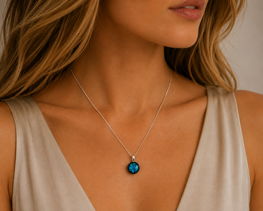 A close-up of a woman wearing a delicate silver chain necklace with a round, dark blue-green stone pendant, paired with a beige sleeveless top.