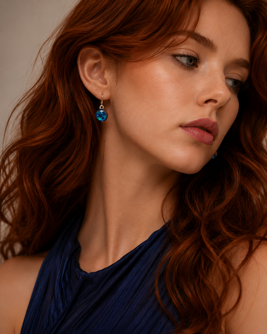 A close-up portrait of a woman with wavy red hair wearing a dark blue halter top and a dangling blue teardrop earring with a silver-toned setting.