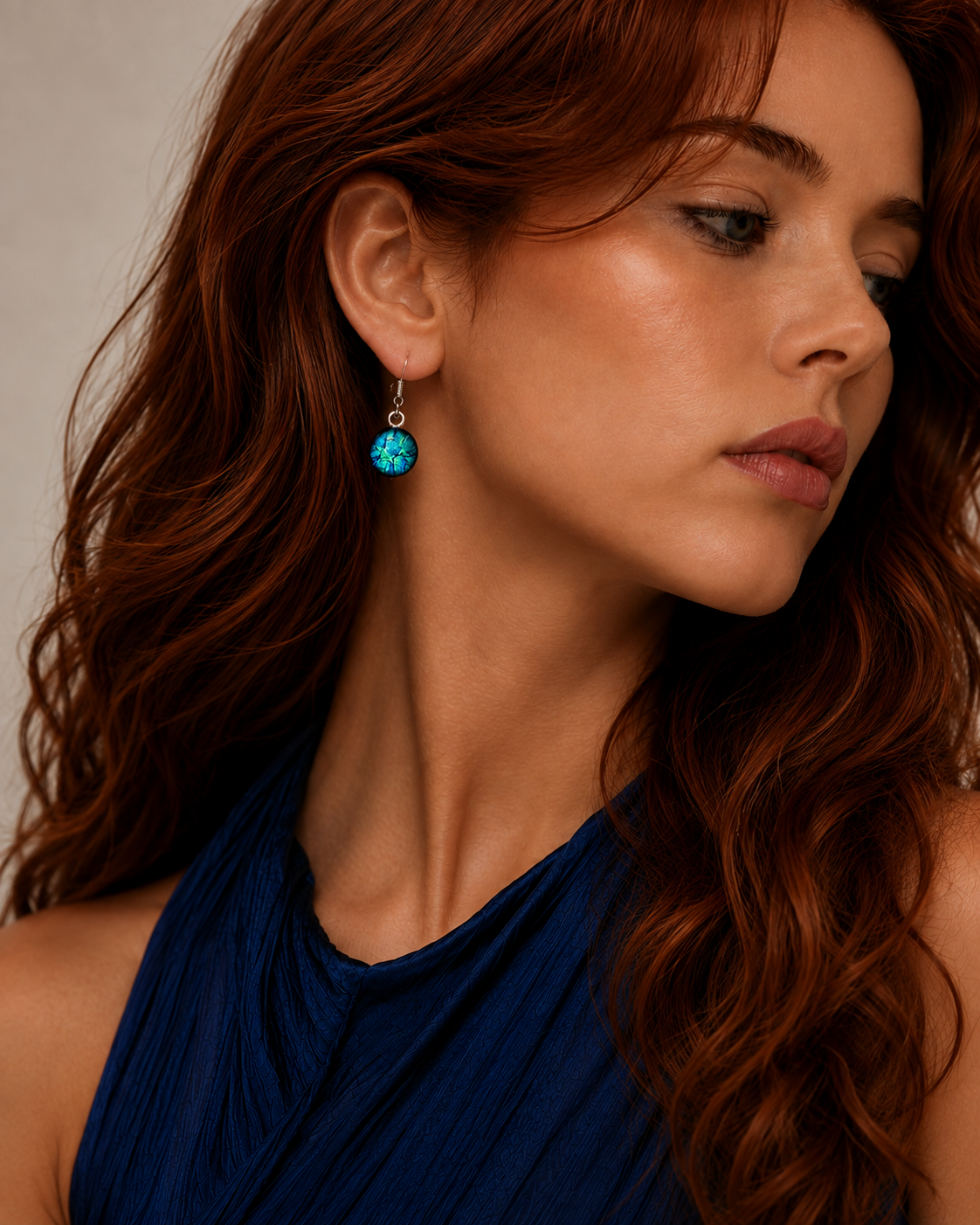 A close-up of a woman with long wavy red hair wearing a small silver hoop earring with a round turquoise-blue stone, paired with a dark blue halter top against a neutral beige background.