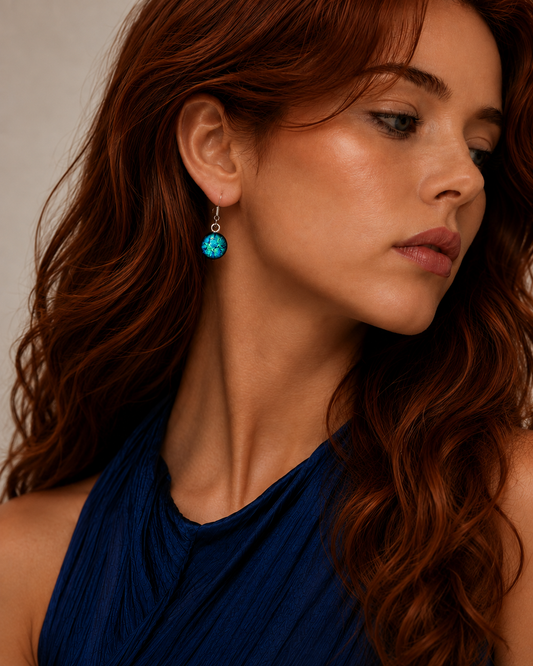 A close-up of a woman with long wavy red hair wearing a small silver hoop earring with a round turquoise-blue stone, paired with a dark blue halter top against a neutral beige background.