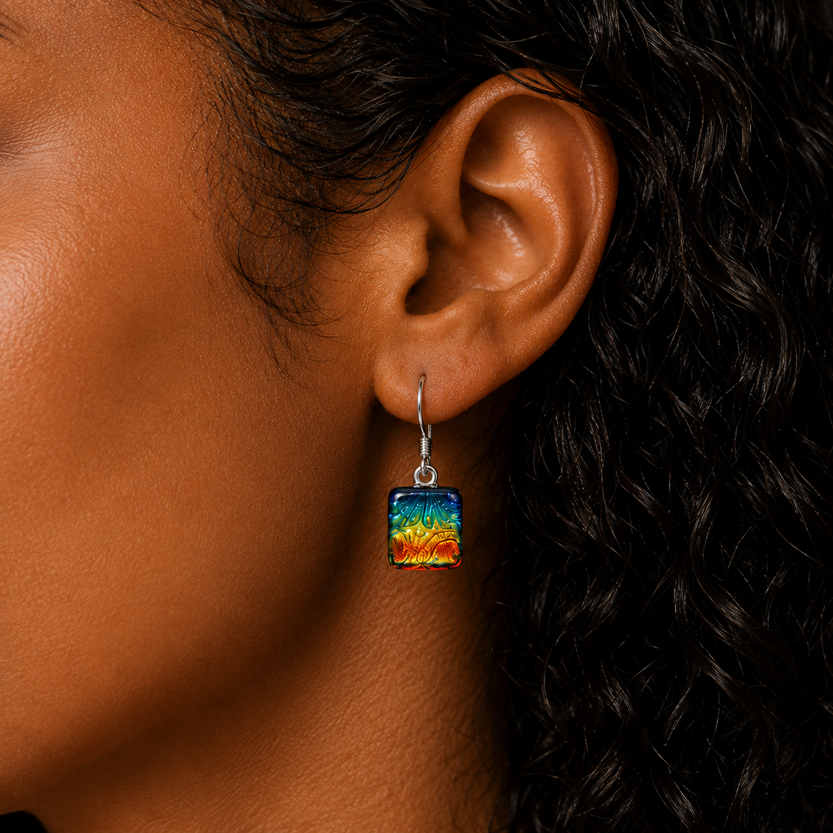 A close-up of a person's ear wearing a square glass drop earring with a vibrant multicolor abstract design in shades of blue, green, yellow, and orange.