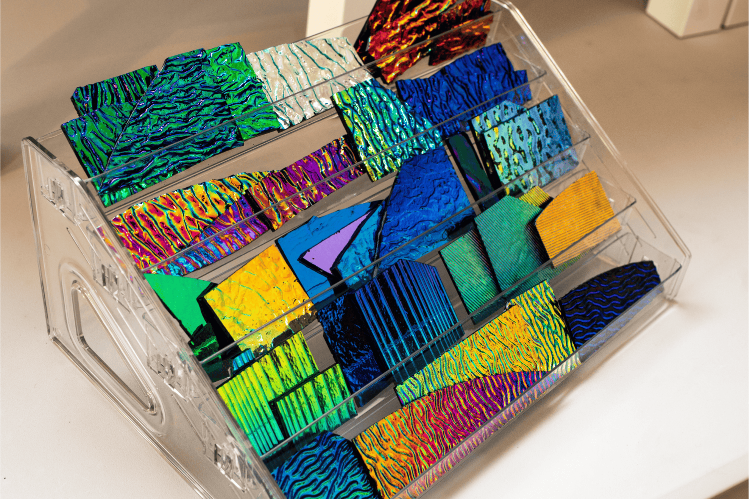 Colourful dichroic glass sheets with textured patterns displayed in a clear organiser, ready for handmade jewellery making.